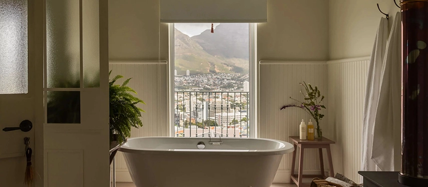 Freestanding bathtub at Dorp Hotel with city views of Cape Town framed by a large window and elegant white panelling.