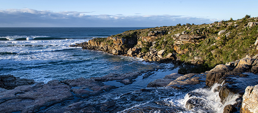 Ocean waves meeting the rocky shoreline under a clear blue sky.