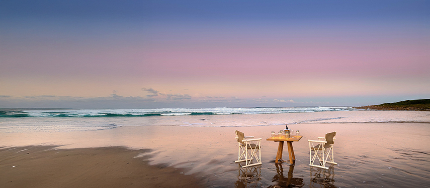 Intimate beach dining setup with table and chairs placed at the water’s edge.