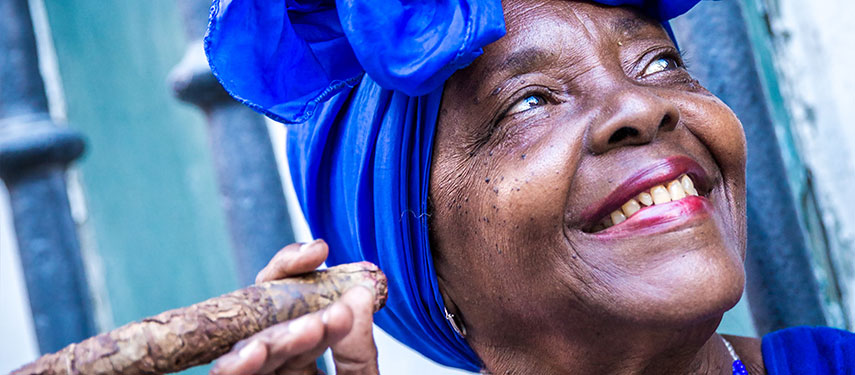 Afro Cuban women in bright blue clothing smiles as she smokes a cigar