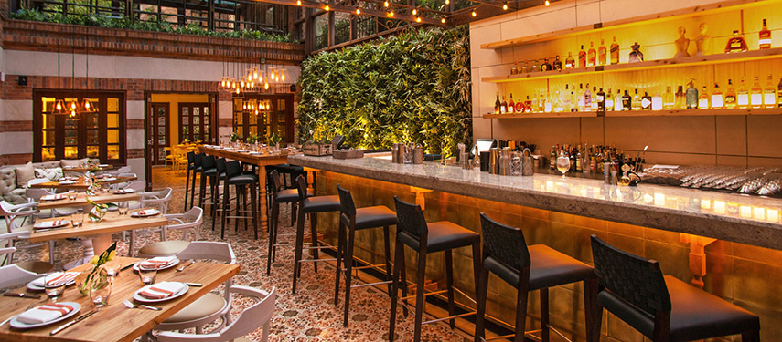 Atmospheric dining and bar area at Four Seasons Casa Medina showcasing a lush green wall, glowing bar, and elegant table settings.