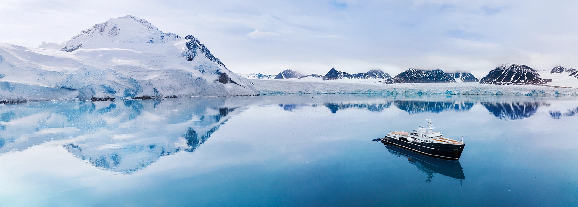 Aqua Lares anchored near a glacier-lined shoreline under a soft grey Arctic sky.
