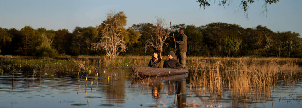 Guests on a traditional mokoro canoe safari at sunset, paddling through reeds in the Okavango Delta with a local guide.