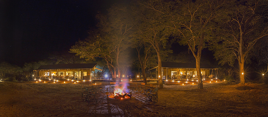 Panoramic night view of Verney’s Camp with firelit seating and tents glowing warmly beneath tall trees.