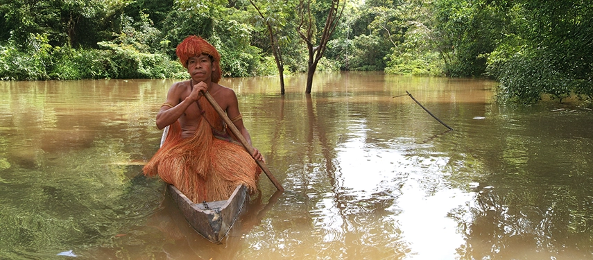 A Boras villager dressed in traditional palm-fibre attire paddling a wooden canoe through flooded forest waters.