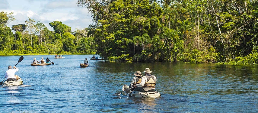 Travellers paddling through a wide Amazon tributary surrounded by dense tropical forest and bright blue skies.