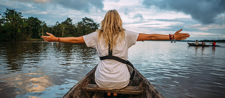A woman seated at the bow of a canoe with arms outstretched, embracing the serenity of an Amazon river at dusk.