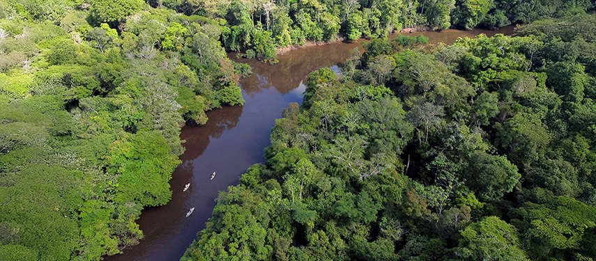 An aerial view of small boats gliding along a winding brown-water river surrounded by dense Amazon rainforest canopy.