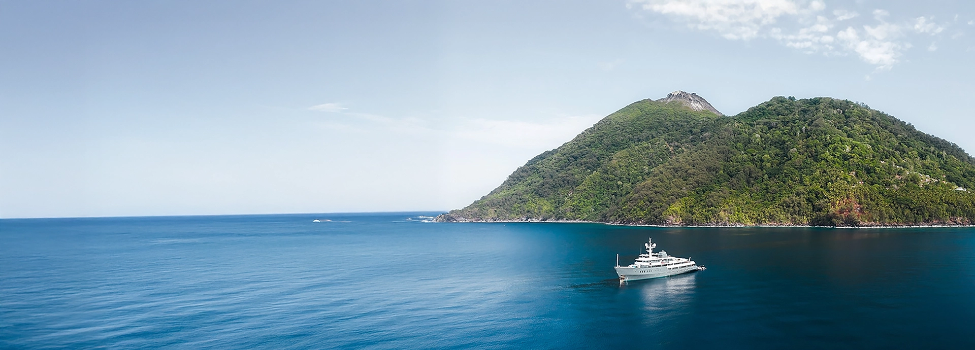 Aqua Blu anchored in tranquil blue seas framed by lush green islands, sunlight glinting on her sleek hull.