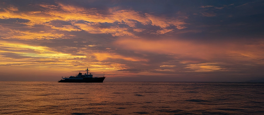 Aqua Blu silhouetted against a radiant sunset sky, glowing clouds mirrored in the still ocean at day’s end.