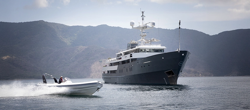 A sleek tender speeds across the water towards Aqua Blu, moored near verdant mountains under soft afternoon light.