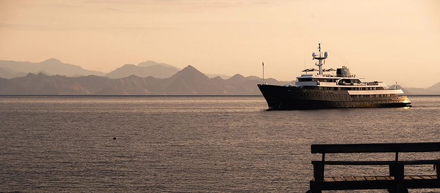 Aqua Blu at anchor under warm golden light, distant mountain silhouettes creating a tranquil Indonesian seascape.