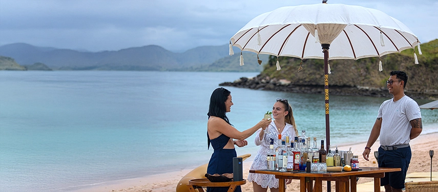 Guests enjoy drinks under white parasols on a secluded beach, with the calm sea stretching toward distant islands.