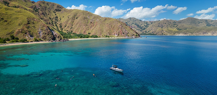A small boat anchors in turquoise waters as snorkellers explore coral gardens near the shores of a lush Indonesian island.