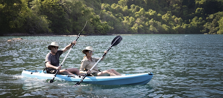 Couple paddling together across calm lagoon waters, framed by dense forest and soft morning sunlight.