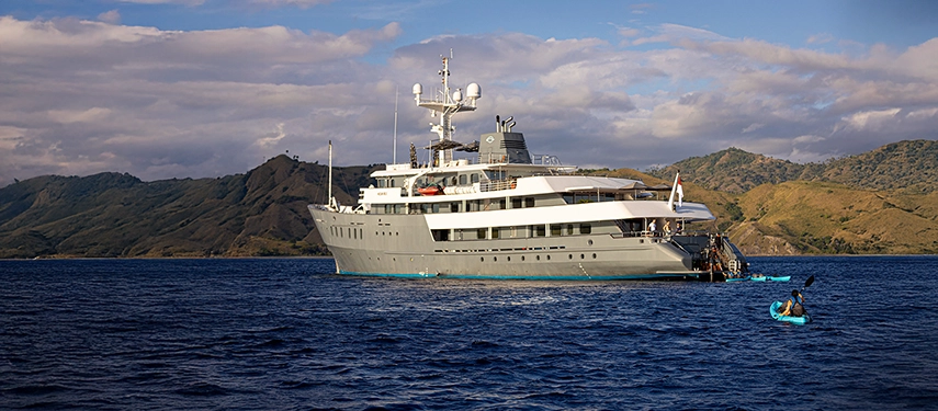 Guest paddling beside Aqua Blu under clear skies, with rugged green islands forming a striking backdrop.