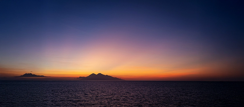 Golden sunset over calm island waters, silhouetting forested hills and casting warm reflections across the sea.