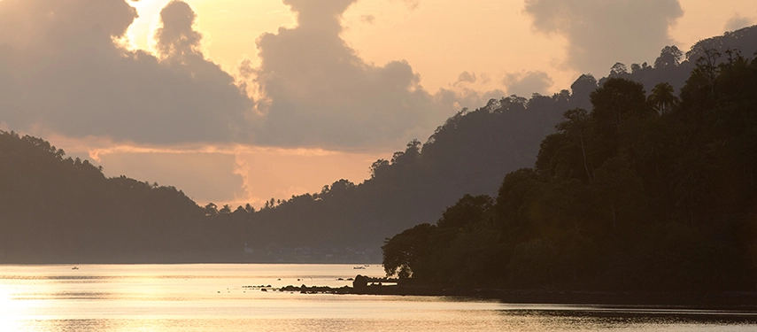 Soft golden light illuminates forested hills and calm reflective waters at dusk, capturing the tranquil beauty of Indonesia’s islands.