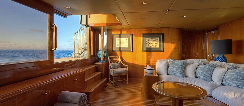 A bright teak-panelled lounge on Aqua Mare opens to sea views, with plush seating, coastal artwork and golden light streaming through wide windows.