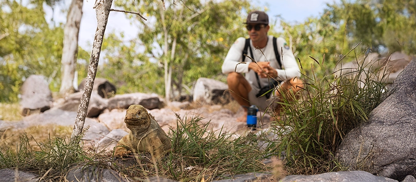 A naturalist crouches near a Galápagos land iguana in dappled shade, observing its calm movements in the island undergrowth.