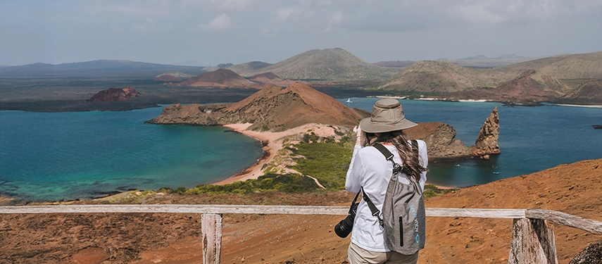 A traveller photographs the sweeping volcanic landscape of Bartolomé Island, with turquoise bays and Pinnacle Rock below.