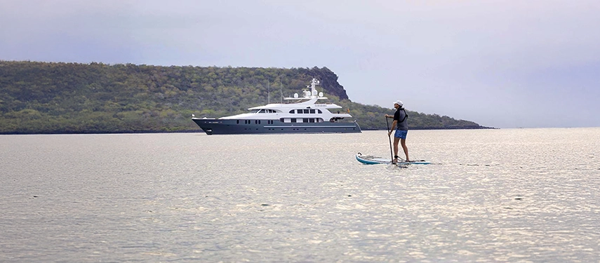 A paddleboarder drifts across calm Galápagos waters with Aqua Mare anchored nearby beneath misty island cliffs.
