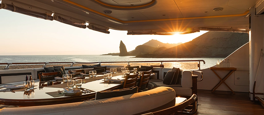 An elegant al fresco dining setup aboard Aqua Mare captures the last rays of sunset over the dramatic coastline of Bartolomé Island.