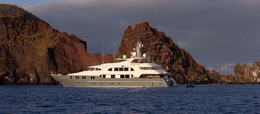 Aqua Mare rests at anchor beside rugged volcanic cliffs, her silver hull glowing softly in the afternoon light.