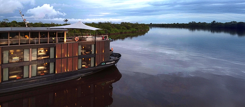 Aqua Nera drifting along the mirrored surface of the Amazon River, surrounded by lush greenery and evening clouds.