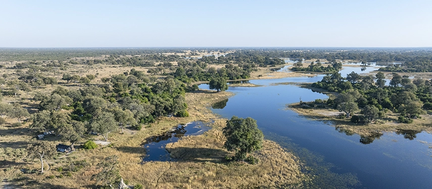 Expansive aerial view of the Okavango Delta showing winding waterways, grassy floodplains, and scattered camp tents.