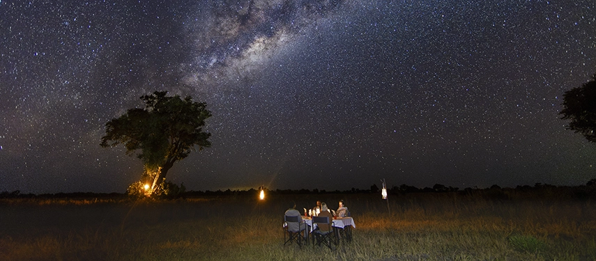 Private dining table set under a star-filled night sky with lanterns glowing around Kweene River Camp.