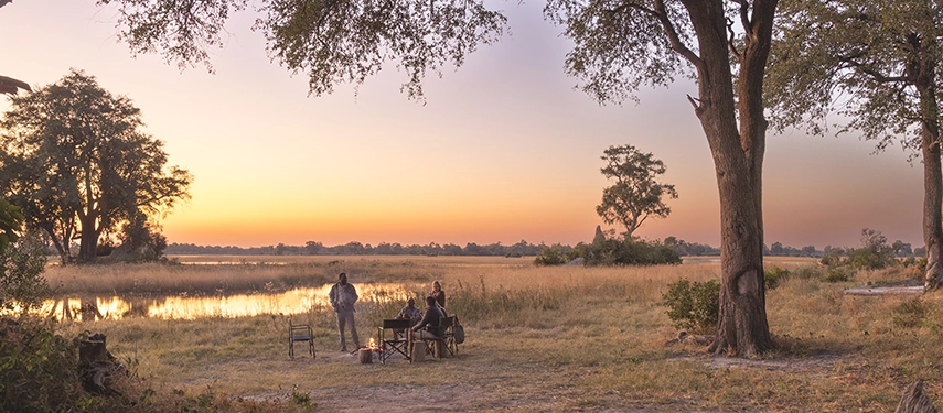 Group of safari travellers positioned around a campfire beside the water at Kweene River Camp during sunset.