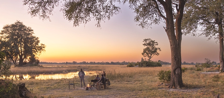 Evening drinks are enjoyed around a crackling campfire at Kweene Camp, set against open floodplains and a glowing sunset.