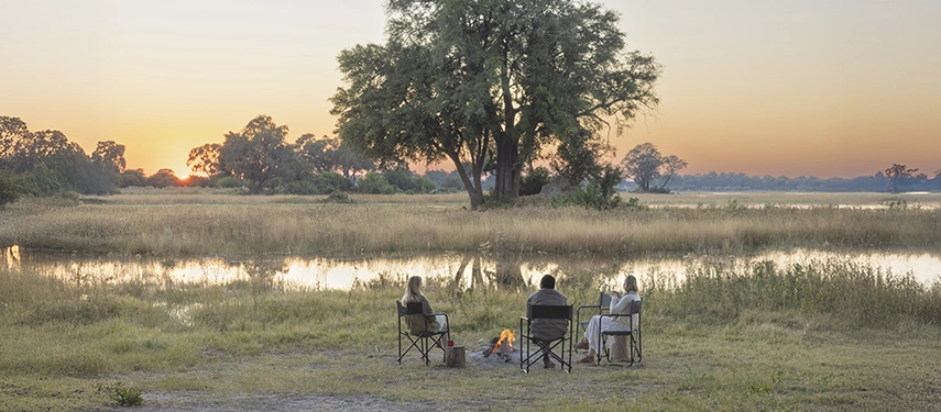 Group of safari goers positioned around a campfire beside the water at Kweene River Camp during sunset.