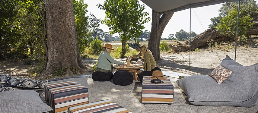Two guests playing a board game on low tables in the shaded lounge tent overlooking the river.