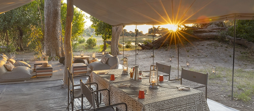 Lantern-lit dining table set beneath a canvas mess tent with lounge seating at sunset in the Delta.