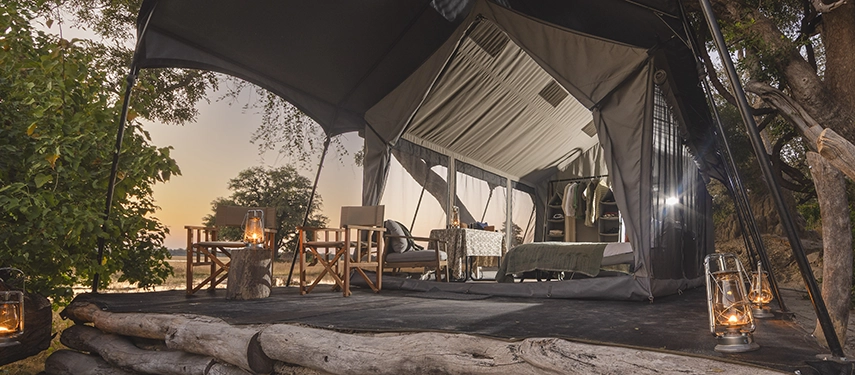 Canvas safari tent with lanterns glowing on a wooden deck, overlooking the Okavango Delta at dusk.