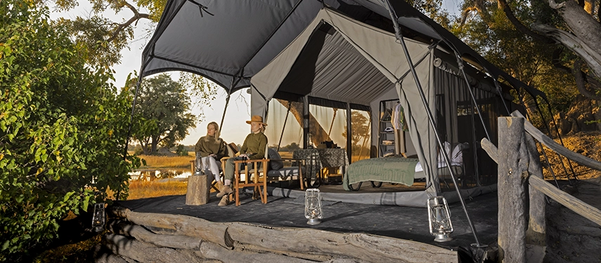 Couple seated on a wooden deck outside their safari tent, overlooking the Delta at golden hour.
