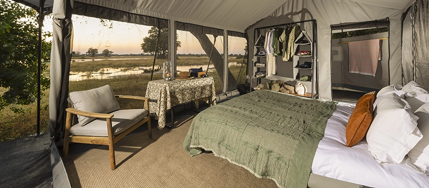 Canvas safari tent interior with double bed, open storage shelves, and a writing table facing Delta views.