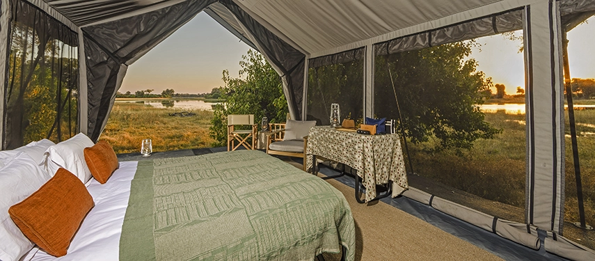 Safari tent bedroom with green bedspread, desk, and wide mesh windows framing the Okavango Delta.