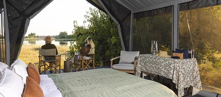 Sunlit safari tent bedroom with king bed, rustic furnishings, and panoramic mesh windows facing the Delta.