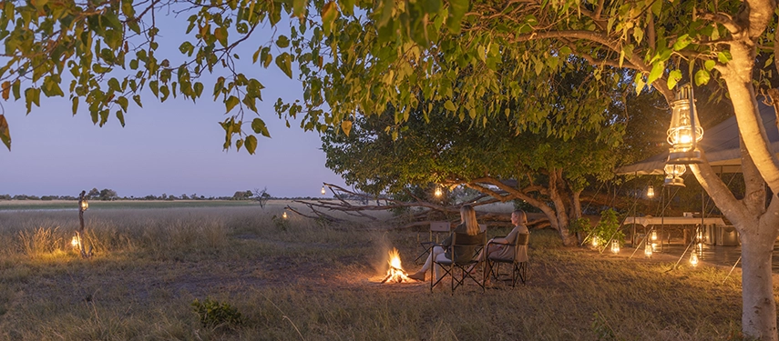 Guests relaxing by the campfire at Magwegwe Camp as lanterns illuminate the bush.