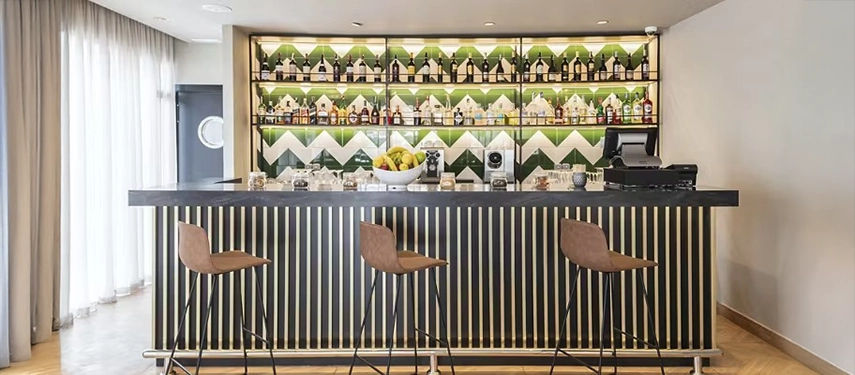 Contemporary bar with striped counter, high stools, and colourful bottles displayed against a patterned green and white wall.