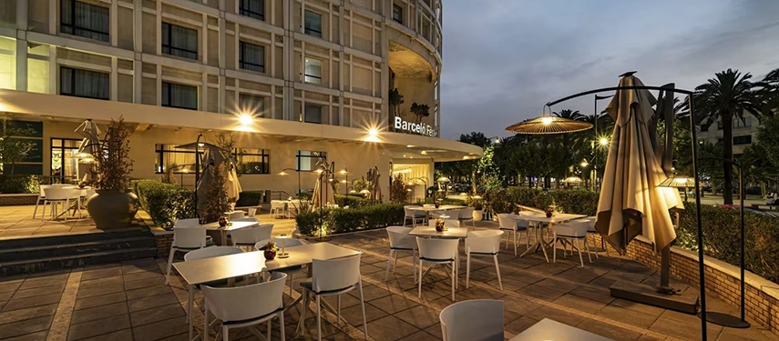 Evening view of the hotel’s outdoor terrace with white tables, soft lighting, and umbrellas beside the modern façade.