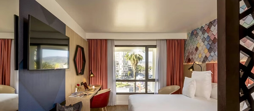 Bright hotel room with city views, a large bed, red curtains, and a desk beneath a wall-mounted TV.
