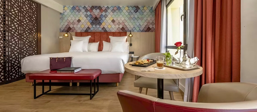 Modern hotel room with king-size bed, red accents, and a small table set with Moroccan tea and pastries.