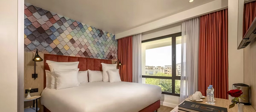 Spacious hotel room with a large bed, colourful tiled headboard design, and wide window letting in natural light.