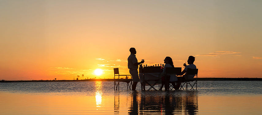 Guests enjoy sundowner drinks with a guide at a table set in the Makgadikgadi salt pans at sunset.