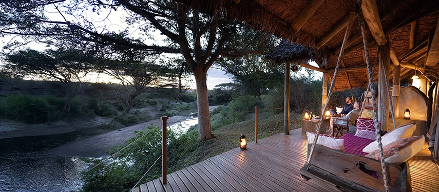 Couple relaxing on a candlelit deck overlooking a riverbank at dusk, enjoying a romantic evening at Basecamp Masai Mara.