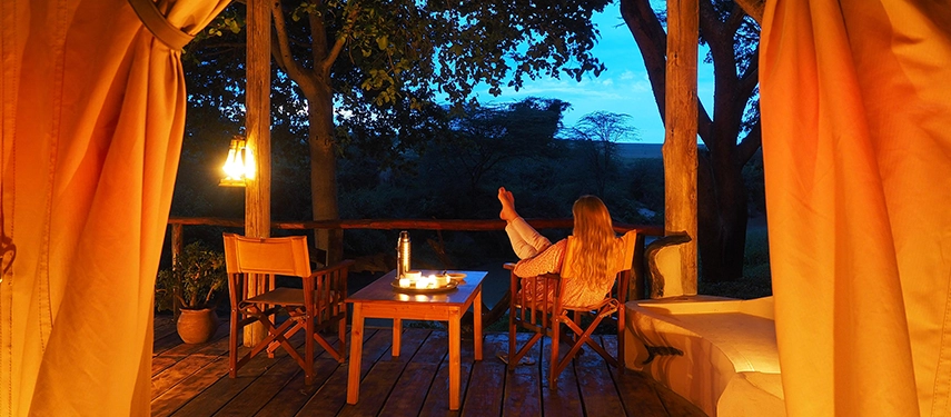 Woman relaxing at dusk on a candlelit veranda at Basecamp Masai Mara, gazing into the wilderness from a cosy safari tent.